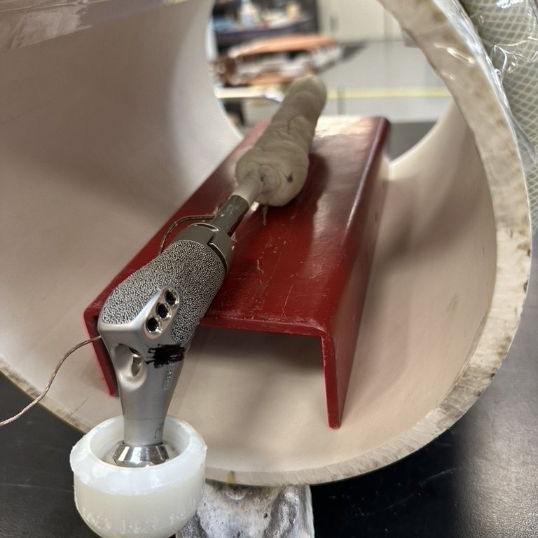 A cemented hip stem resting on a red bench fixture inside a large plastic tube, with a thermocouple wire attached to the implant.